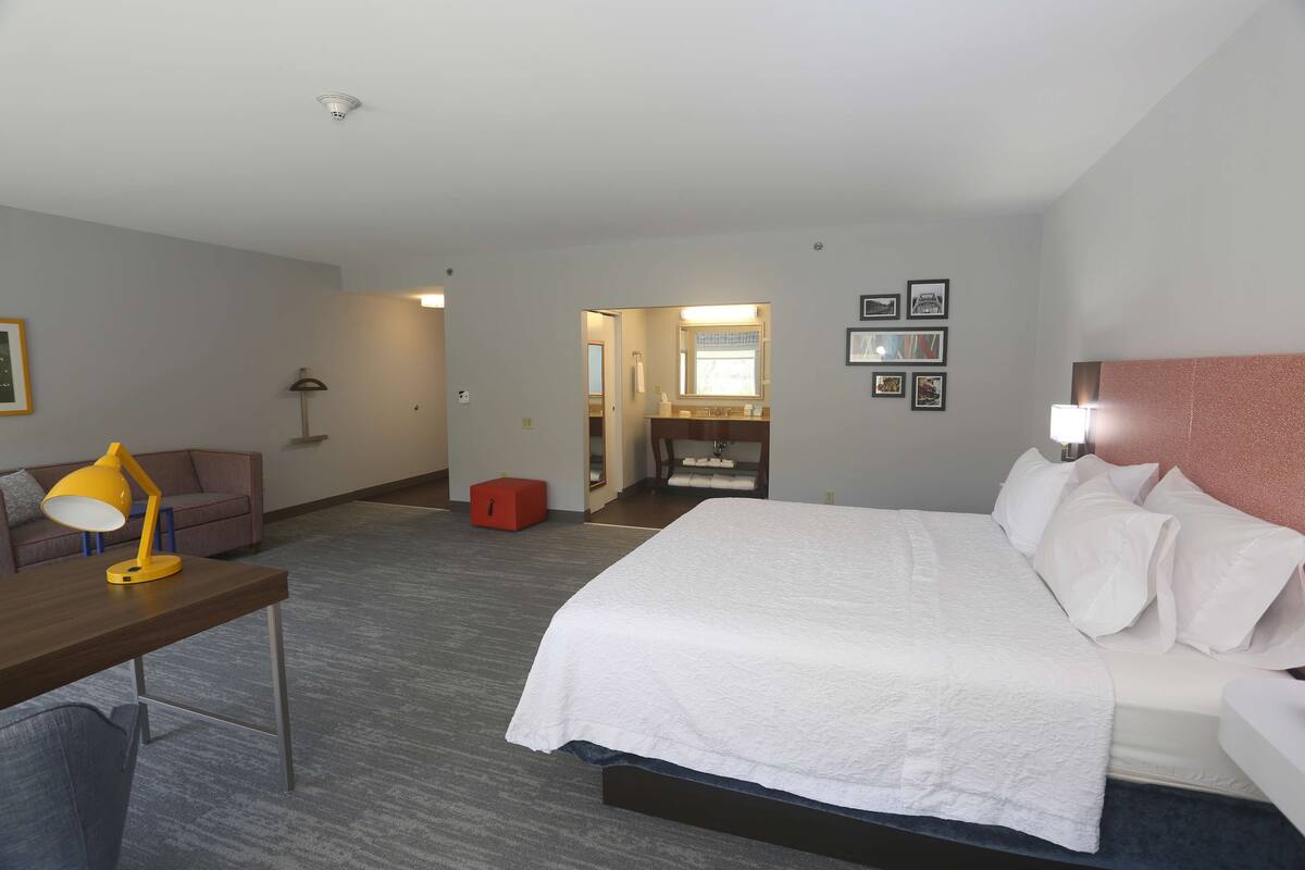 A hotel room with a bed, a desk with a lamp, a sofa, a bathroom with a mirror, and framed pictures on the wall.