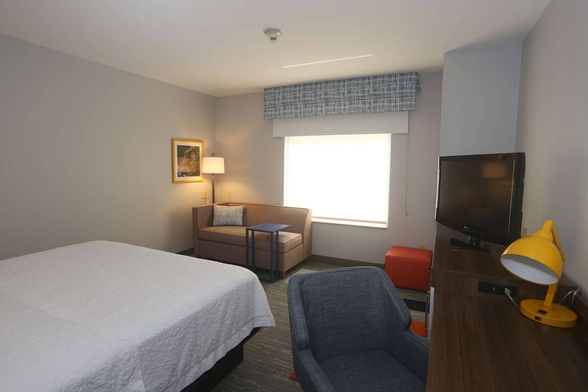 A hotel room with a bed, a sofa, a TV, a desk with a lamp, and a window with a view.