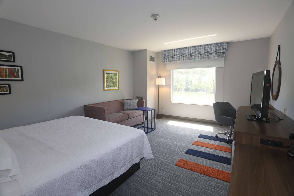 A hotel room with a bed, a desk, a chair, a sofa, a lamp, and a window with a view of trees.