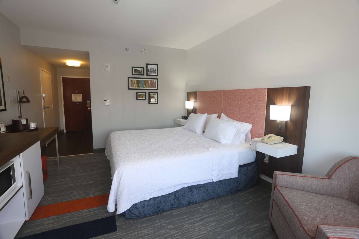 A hotel room with a large bed, a desk, a chair, a nightstand with a lamp, and a wall-mounted phone.