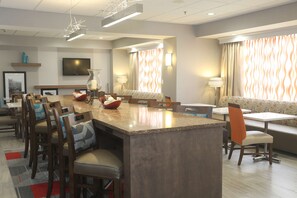 A modern hotel dining area with a central island, seating arrangements, and a television on the wall.
