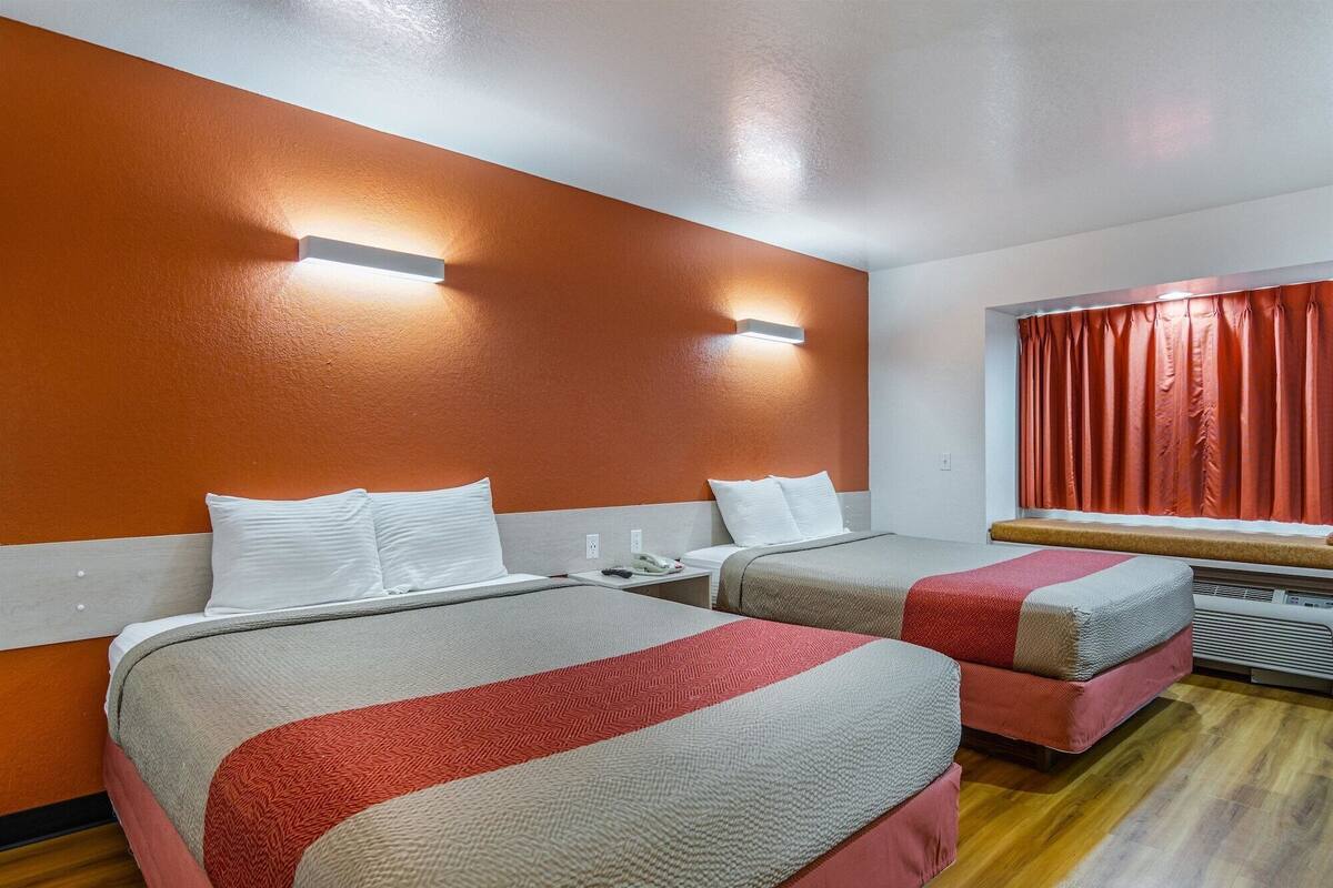 A hotel room with two beds, a window with red curtains, and a wall-mounted air conditioning unit.