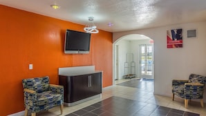 A reception area with orange walls, a flat-screen TV mounted above a dark-colored cabinet, and two patterned chairs. - Motel 6 McAlester, OK (Krebs)