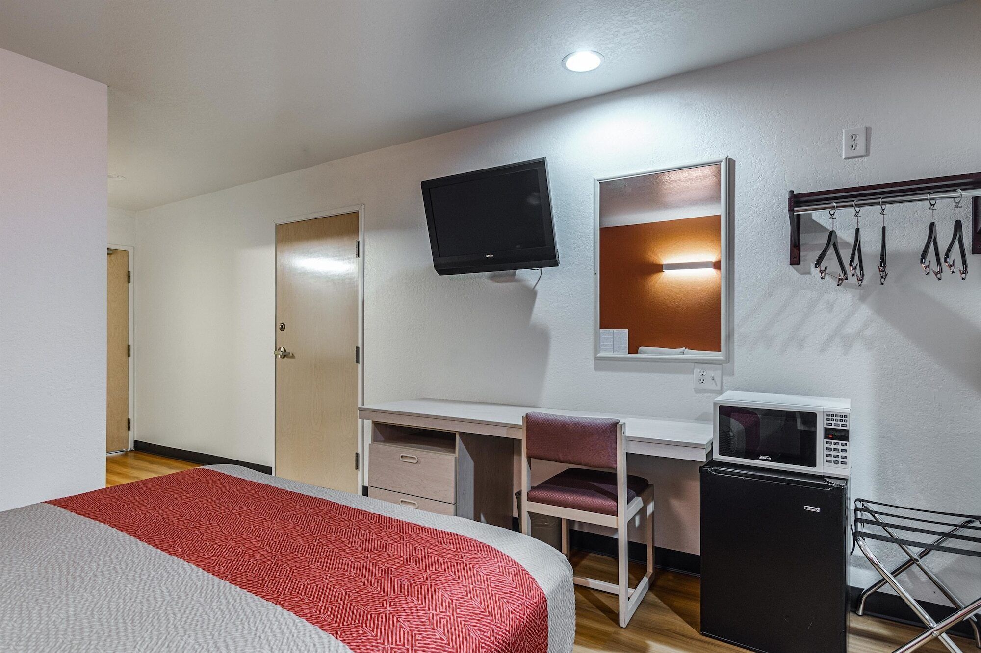 Quarto de hotel com uma cama, escrivaninha, cadeira, televisão, micro-ondas e um pequeno frigorífico.