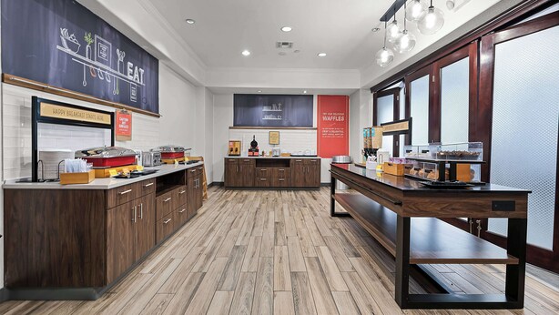 A modern kitchen with wooden cabinets, a central island, and a red banner advertising waffles. - Hampton Inn Seguin (Seguin)