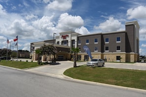 A Hampton Inn hotel with a modern design, featuring a large entrance, a parking area, and a clear sky with clouds.