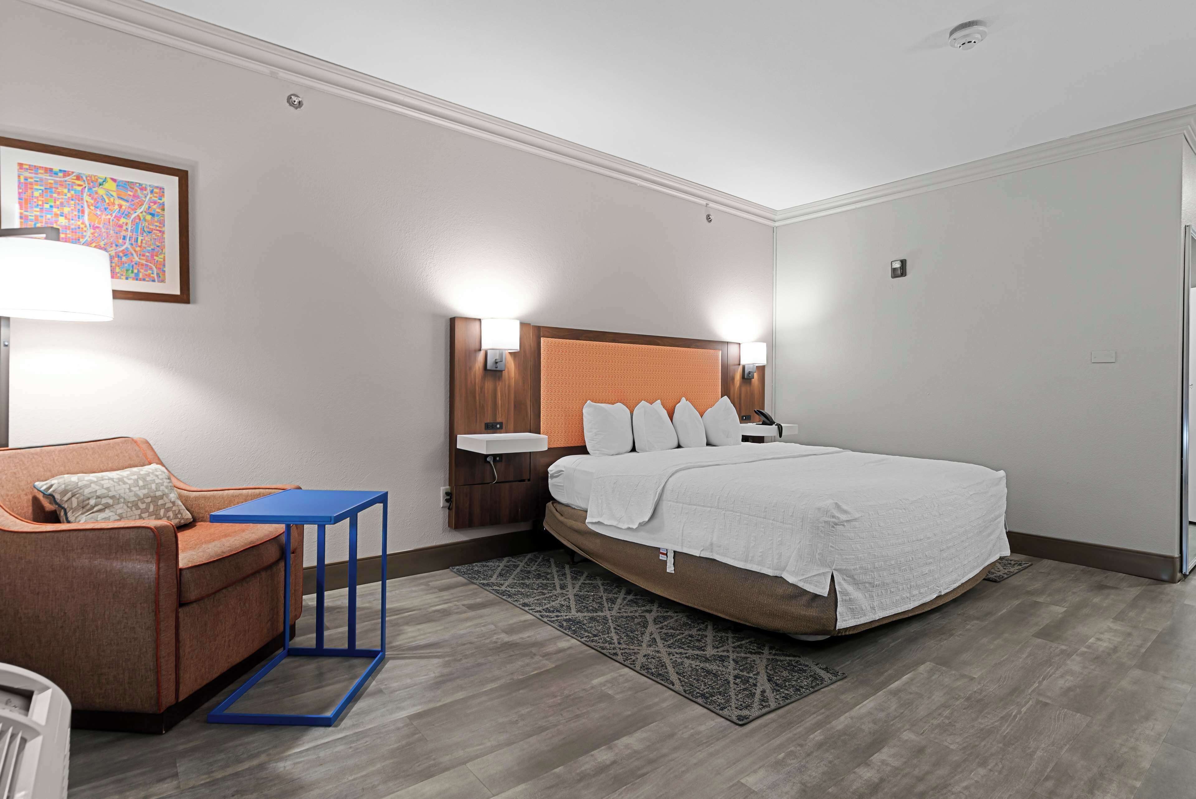 Une chambre d’hôtel comprenant un lit, une chaise, une petite table, une table de chevet, une étagère fixée au mur et un tapis à motifs.
