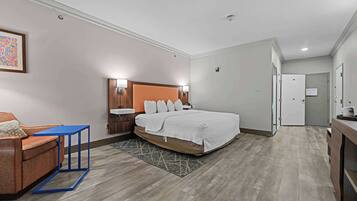 A modern hotel room with a bed, a small table, a sofa, and a bathroom visible through an open door.