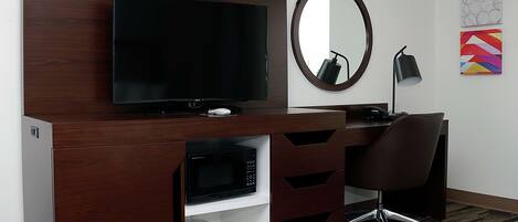 A modern hotel room with a dark wood desk, a flat-screen TV, a round mirror, a black desk lamp, and a chair.