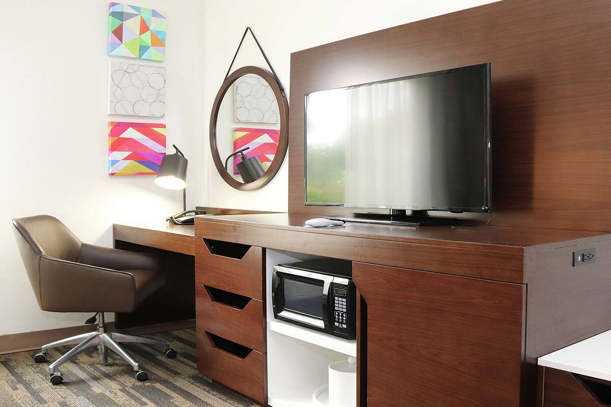 Habitación de hotel con un escritorio de madera, televisión de pantalla plana, microondas, una silla y una obra de arte en la pared.
