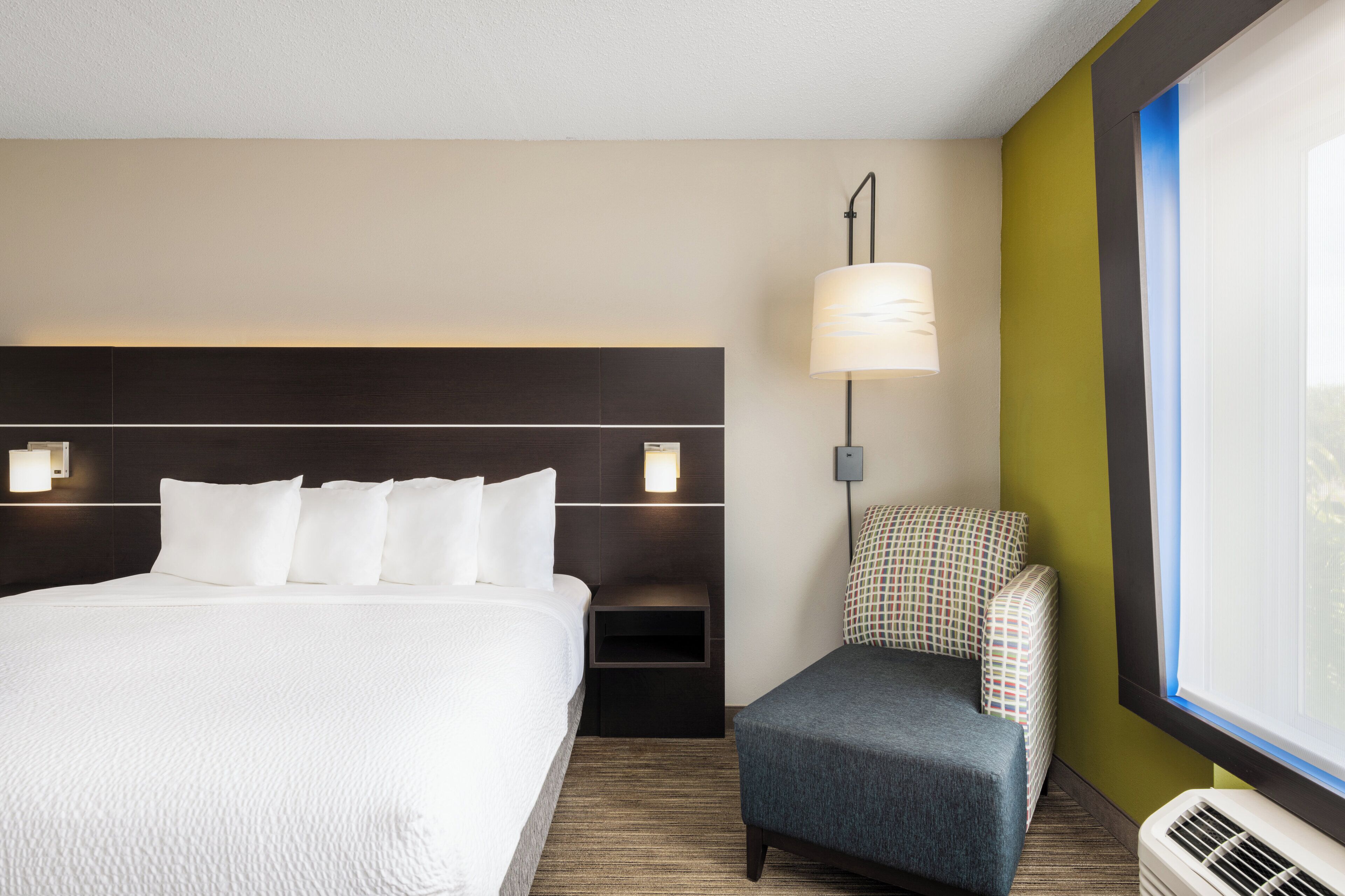 Ein Hotelzimmer mit einem großen Bett, einem Nachttisch mit Lampe, einer kleinen Sitzecke und einem Fenster mit Jalousien.