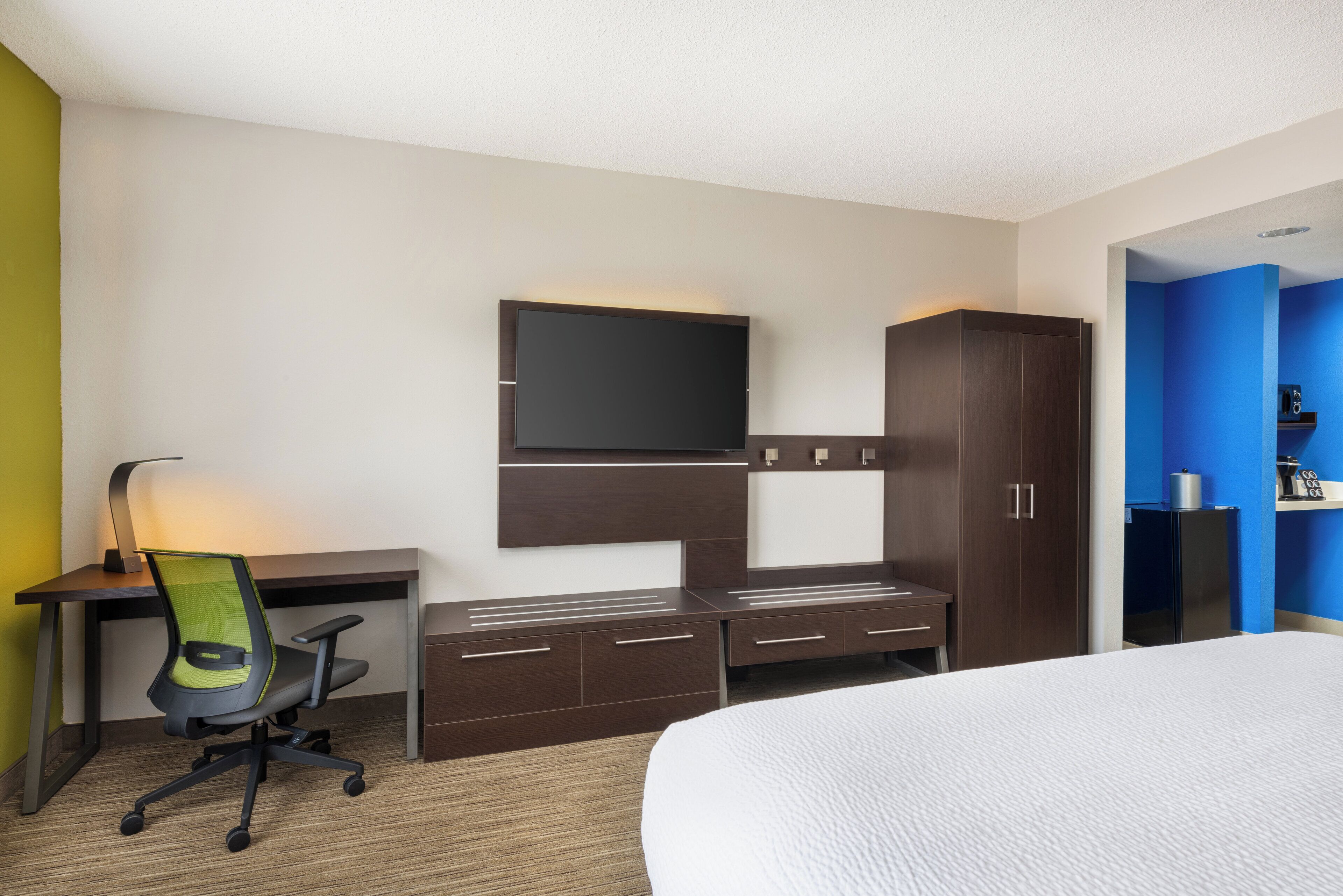 A hotel room with a bed, a desk with a chair, a TV mounted on the wall, and a wardrobe.