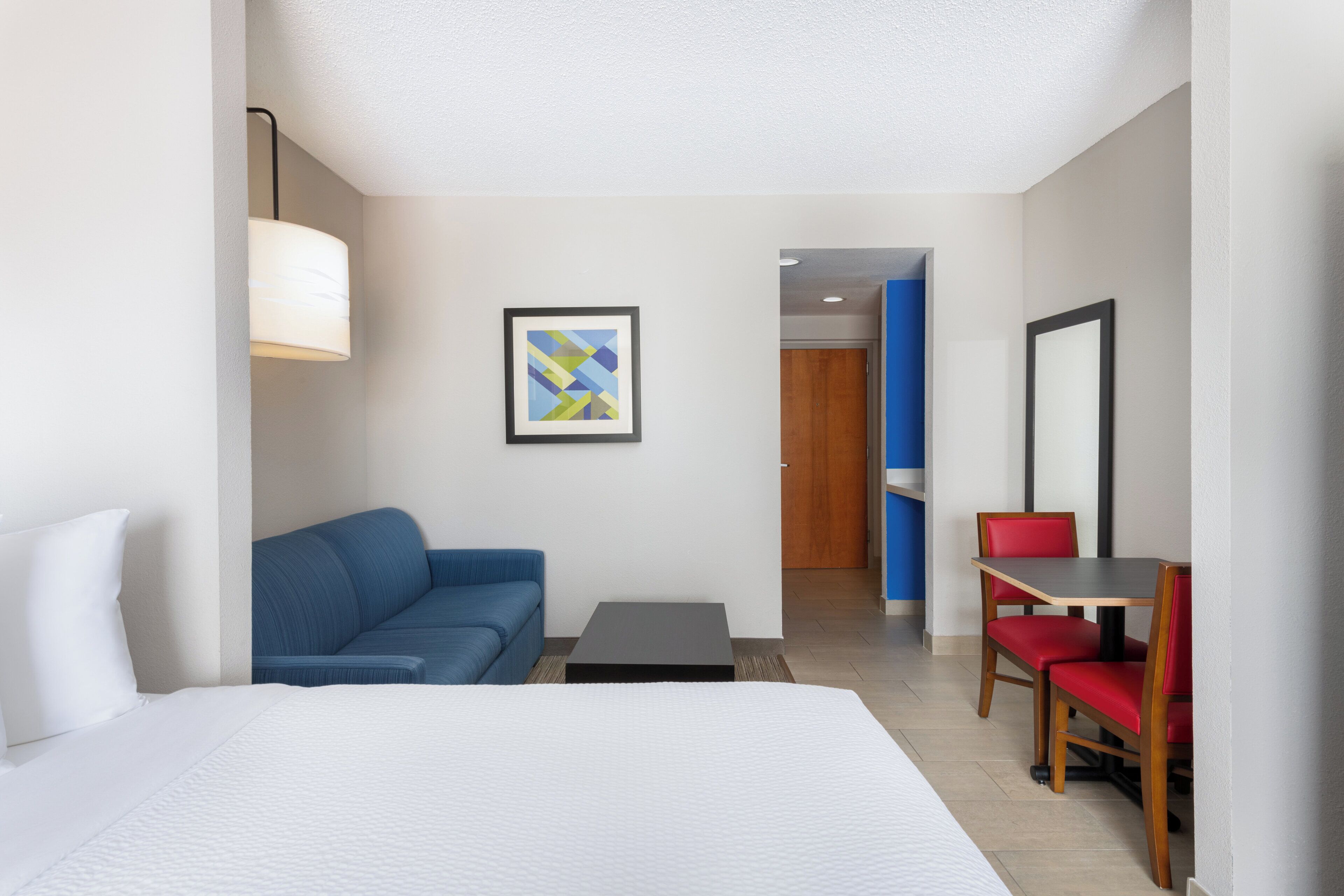 Habitación de hotel con cama, sofá azul, mesita pequeña, silla roja, cuadro enmarcado y puerta que da a otra habitación.