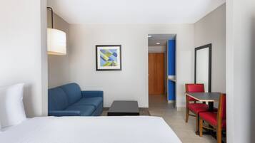 A hotel room with a bed, a blue sofa, a small table, a red chair, a framed picture, and a door leading to another room.