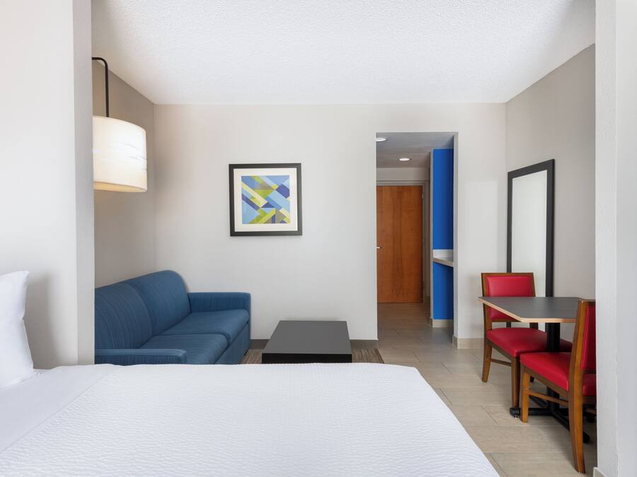 A hotel room with a bed, a blue sofa, a small table, a red chair, a framed picture, and a door leading to another room.