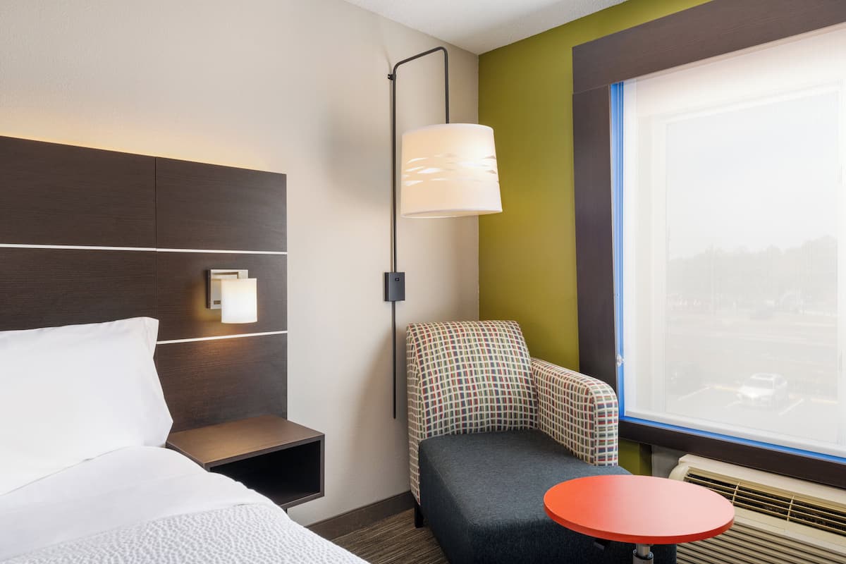 A hotel room with a bed, a small table, a chair, a lamp, and a window with a city view.