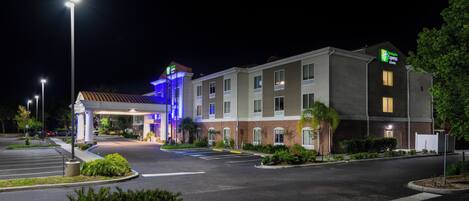 A hotel with a well-lit entrance, a parking lot, and a landscaped area with bushes.