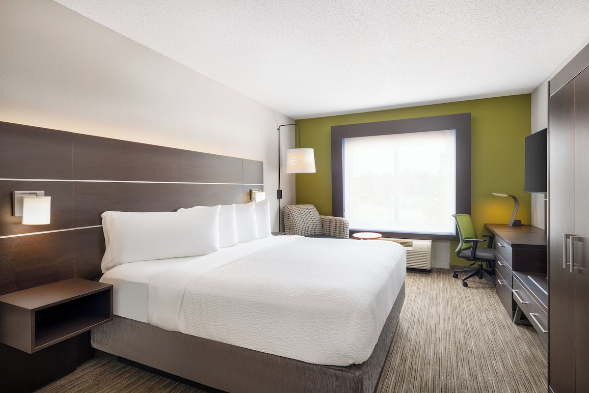 A hotel room with a large bed, a desk, a chair, a TV, and a window with blinds.