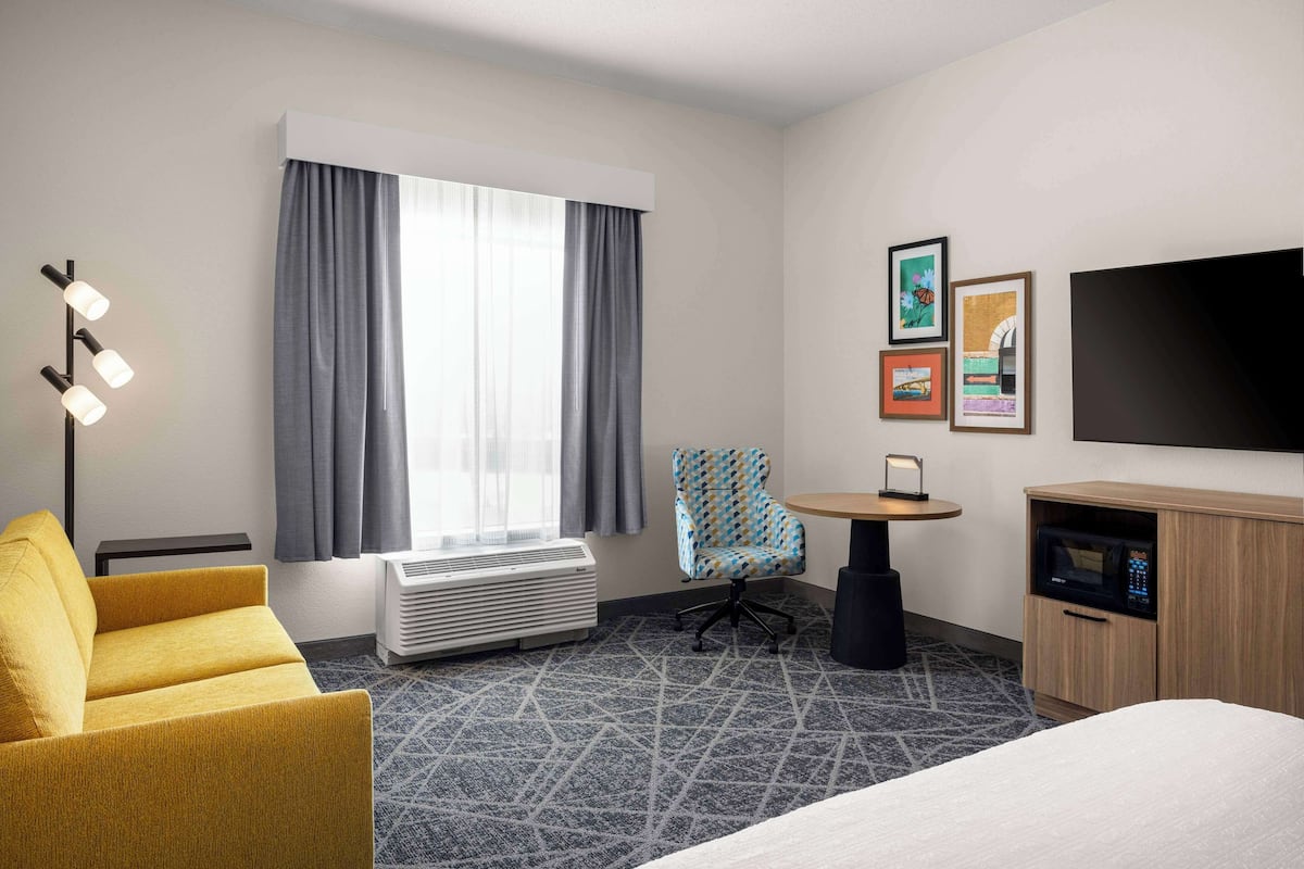 A hotel room with a bed, a yellow armchair, a desk with a chair, a flat-screen TV, a microwave, and a window with curtains.