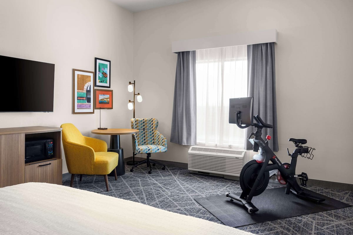 A hotel room with a bed, a desk, a chair, a TV, a bike, and a window with curtains.
