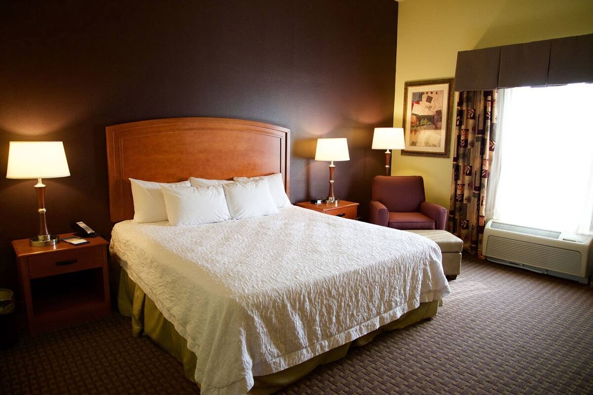 A hotel room with a large bed, two bedside tables with lamps, a chair, a window with curtains, and a wall-mounted air conditioner.