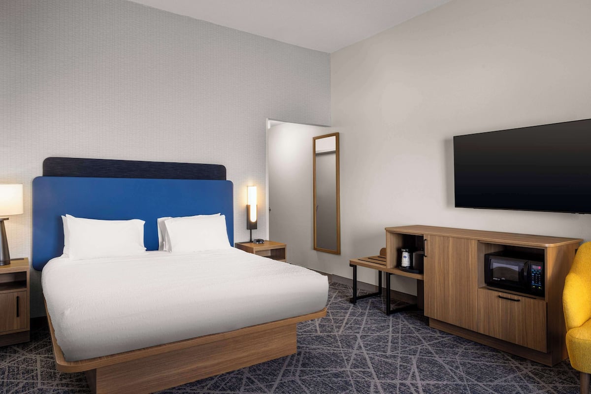 A hotel room with a large bed, a flat-screen TV, a wooden cabinet with a microwave, and a small table.