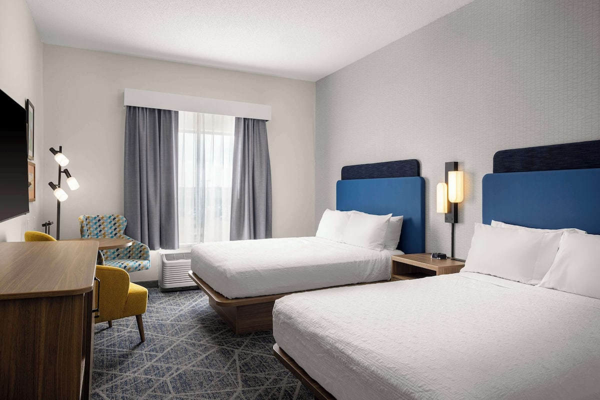 A hotel room with two beds, a desk, a chair, a TV, and a window with curtains.