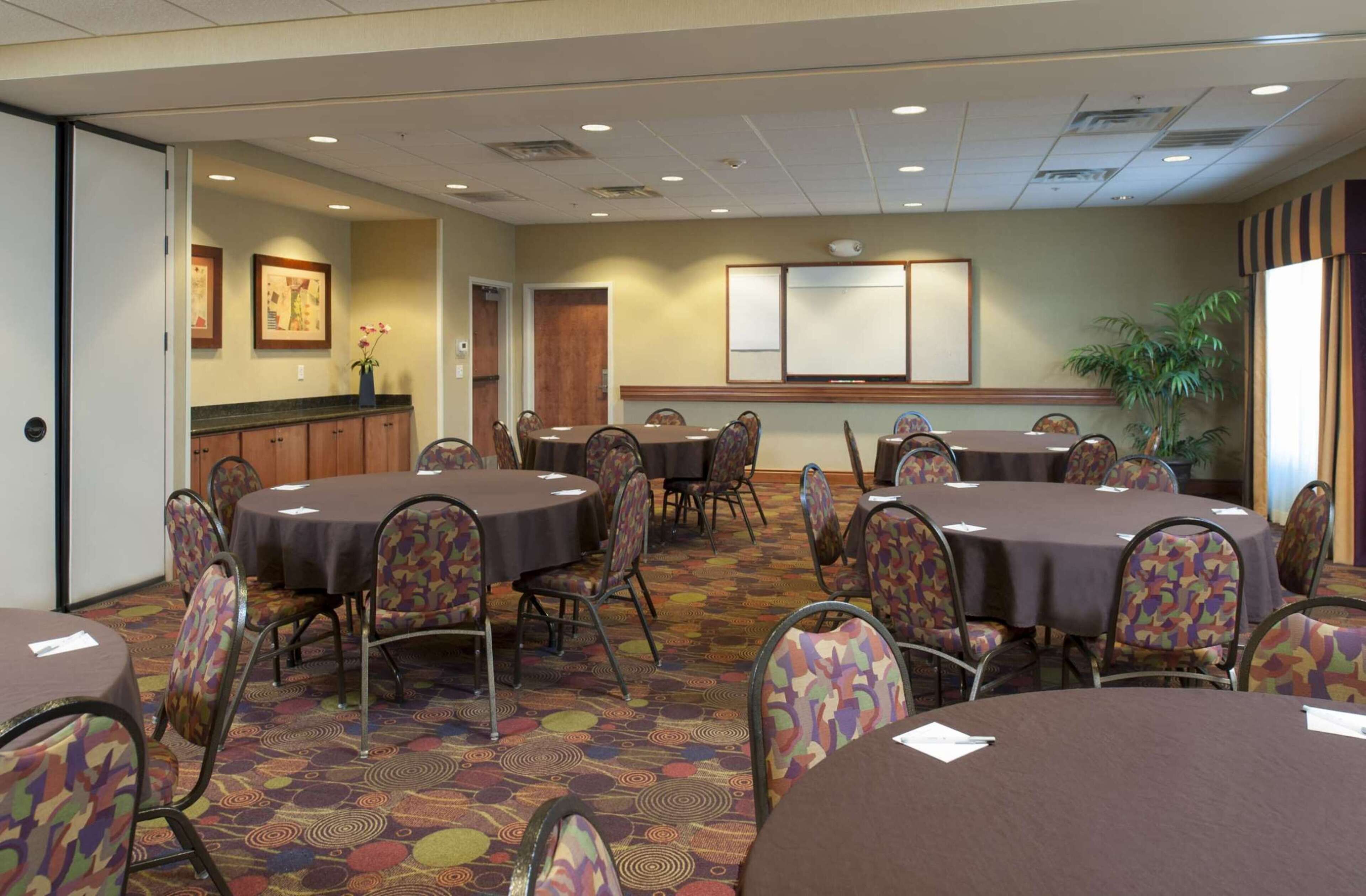 A conference room with round tables and chairs, a projector screen, framed pictures on the wall, and a potted plant.