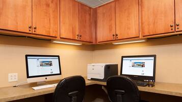 A corner office with two desks, each with a desktop computer, a keyboard, and a mouse. The room has wooden cabinets and a light-colored wall.