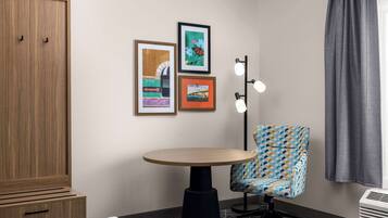 A hotel room with a wooden wardrobe, a round table, a patterned armchair, and three framed artworks on the wall.