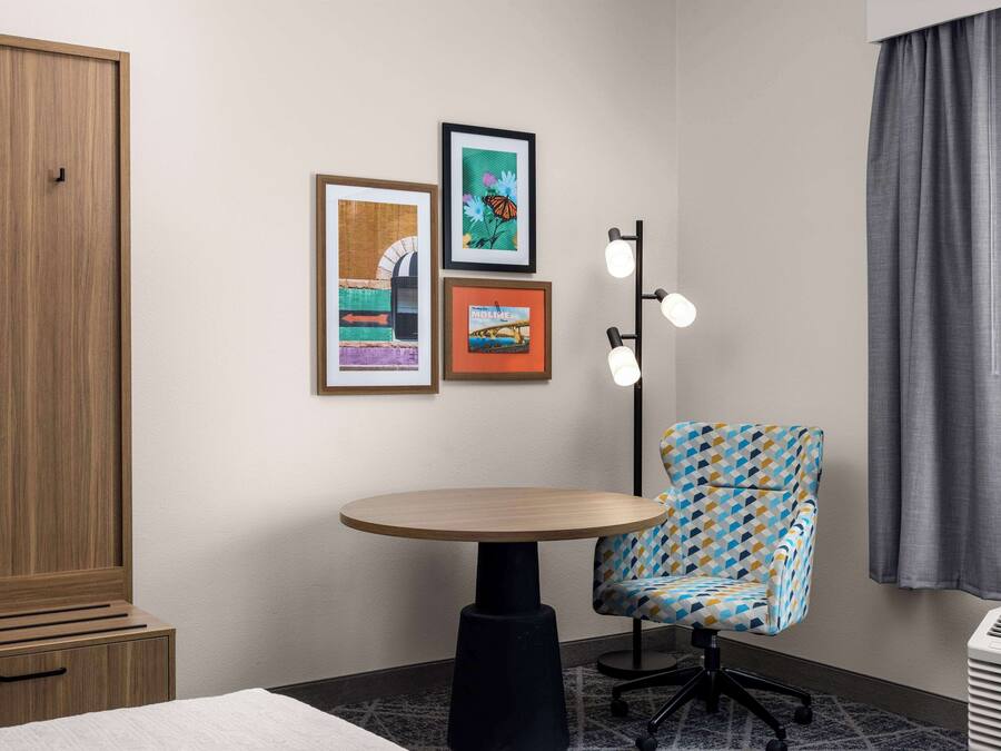 A hotel room with a wooden wardrobe, a round table, a patterned armchair, and three framed artworks on the wall.