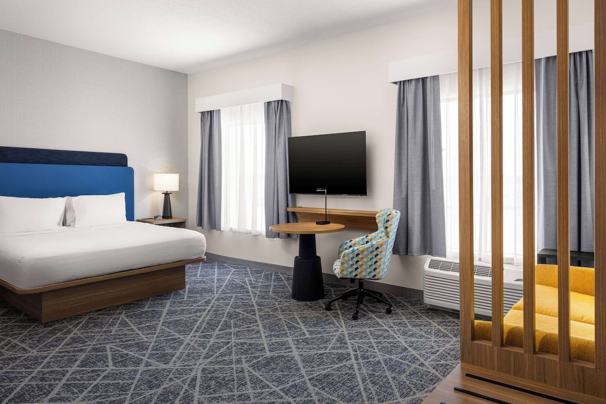 A hotel room with a bed, desk, chair, TV, and air conditioning unit.