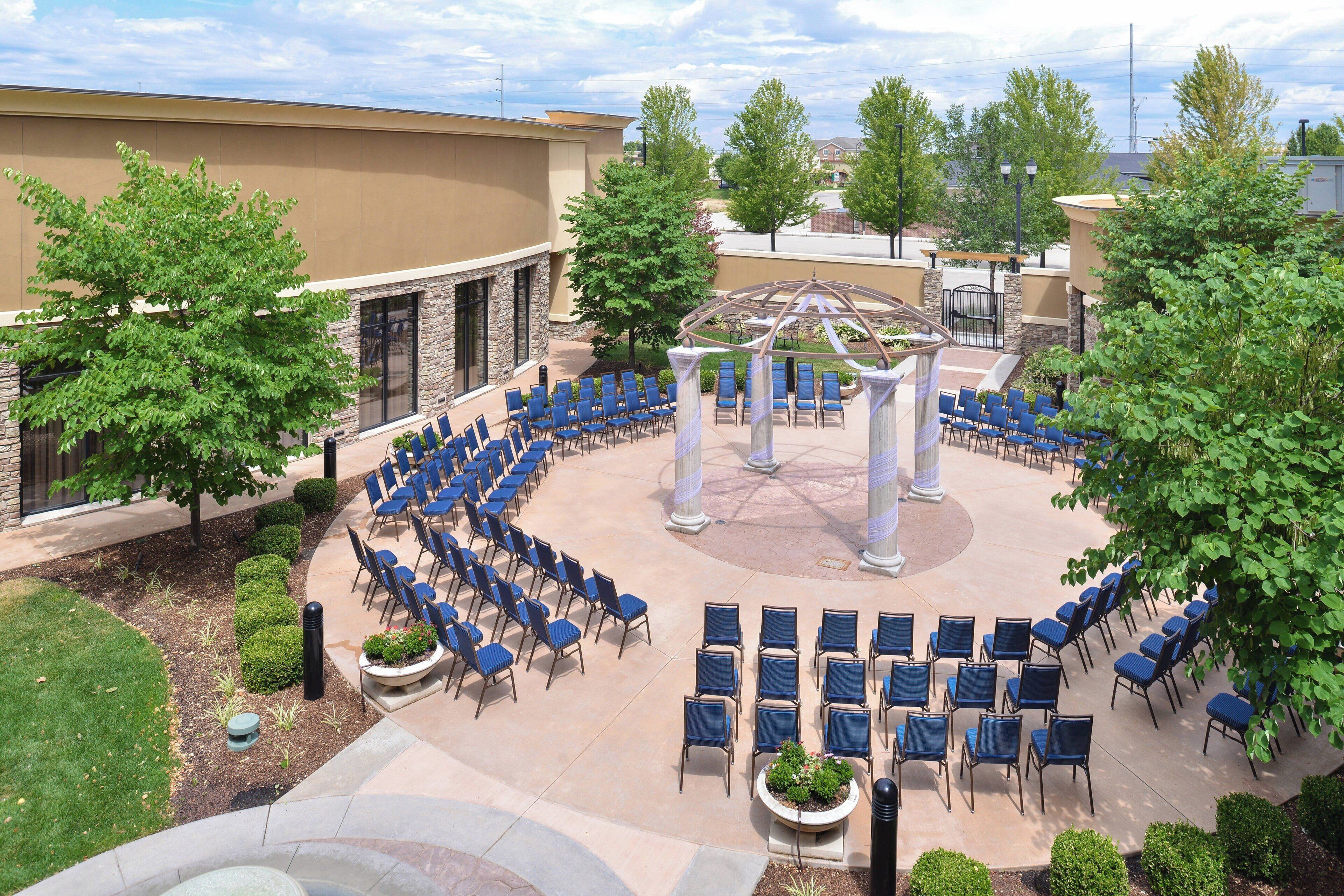 A courtyard with rows of blue chairs arranged in a semi-circle, a central arbor, and a building with stone columns.