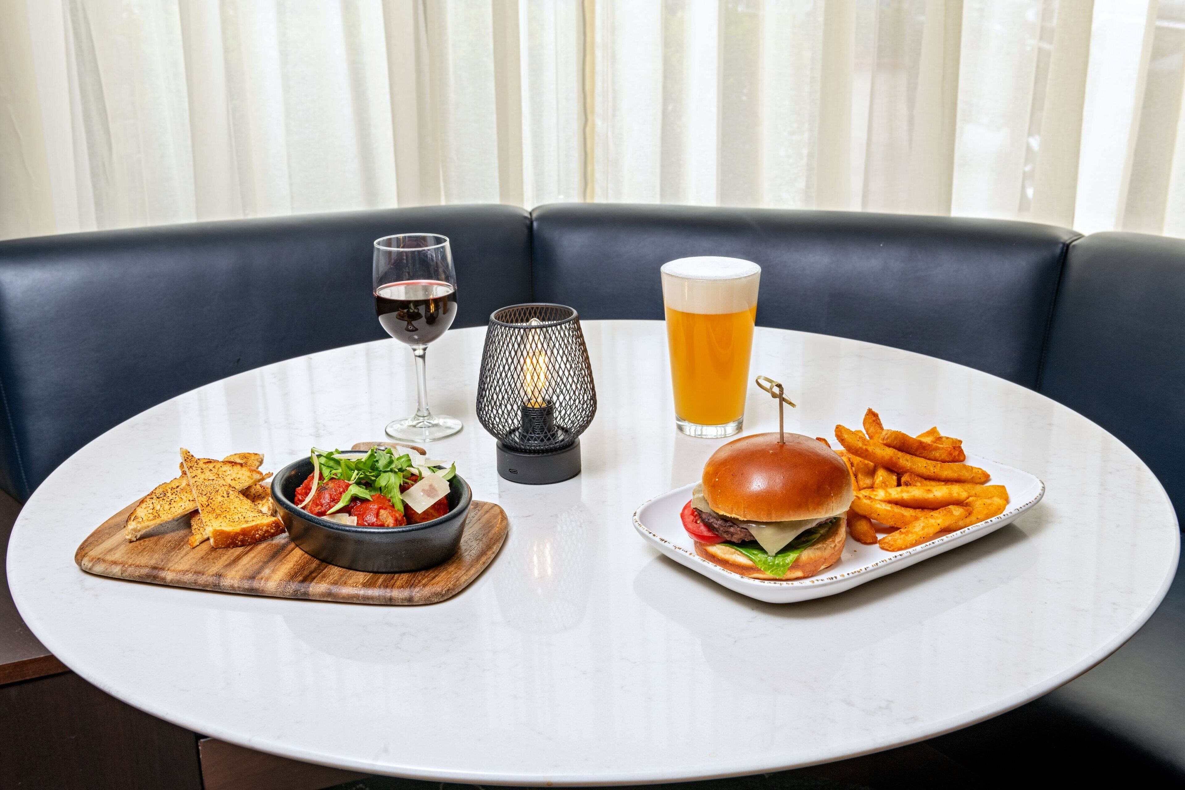 Una mesa redonda de comedor con cubierta de mármol blanco, rodeada por un banco tapizado en azul oscuro. Sobre la mesa, hay un plato con una hamburguesa y papas fritas, un bol de ensalada, una copa de vino tinto y una cerveza.