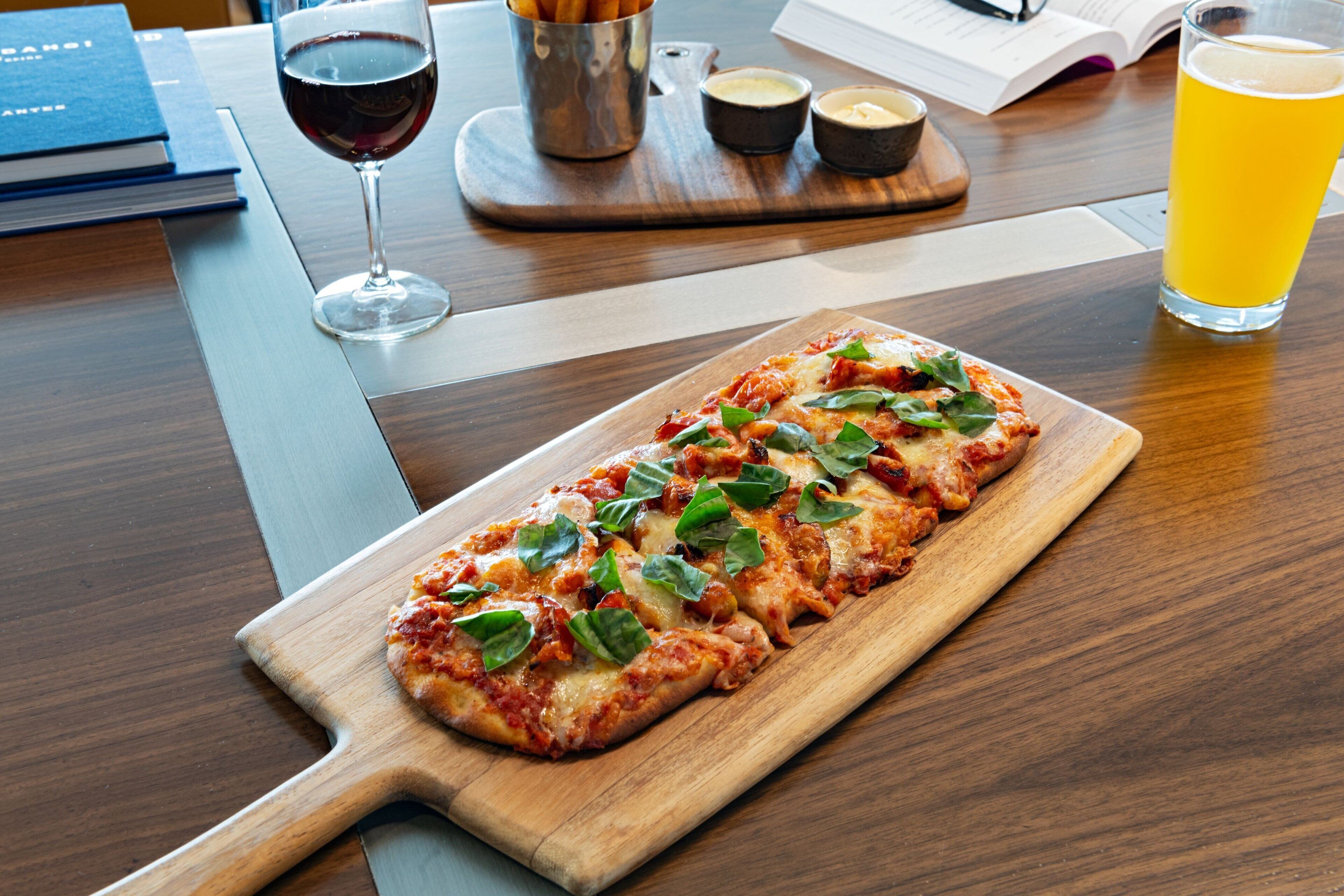 Una tabla de madera con una pizza, una copa de vino tinto y un vaso de jugo de naranja sobre una mesa de madera.