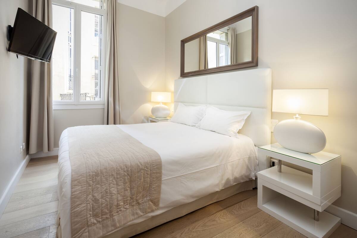 A neatly arranged hotel room with a large bed, a flat-screen TV mounted on the wall, a bedside table with a lamp, and a window with curtains.