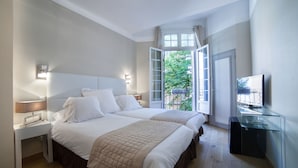 A bedroom with two beds, a TV, and a window with a view of greenery. - Hôtel De France (Aix-en-Provence)