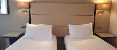 A hotel room with two single beds, each with a pillow and a nightstand. There are wall-mounted lamps on either side of the beds.