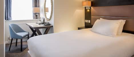 A hotel room with a large bed, a desk with a chair, a lamp, and a wall-mounted light fixture.