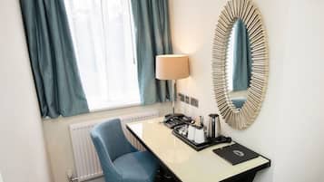A hotel room with a desk, chair, lamp, mirror, and a view of the outside through a window with curtains.