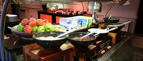 A buffet setup with bowls of cereal, fruits, and a clock on the wall.