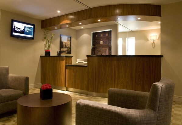 Un área de recepción con un mostrador de madera, un televisor montado en la pared y dos sillones con una pequeña mesa en primer plano.