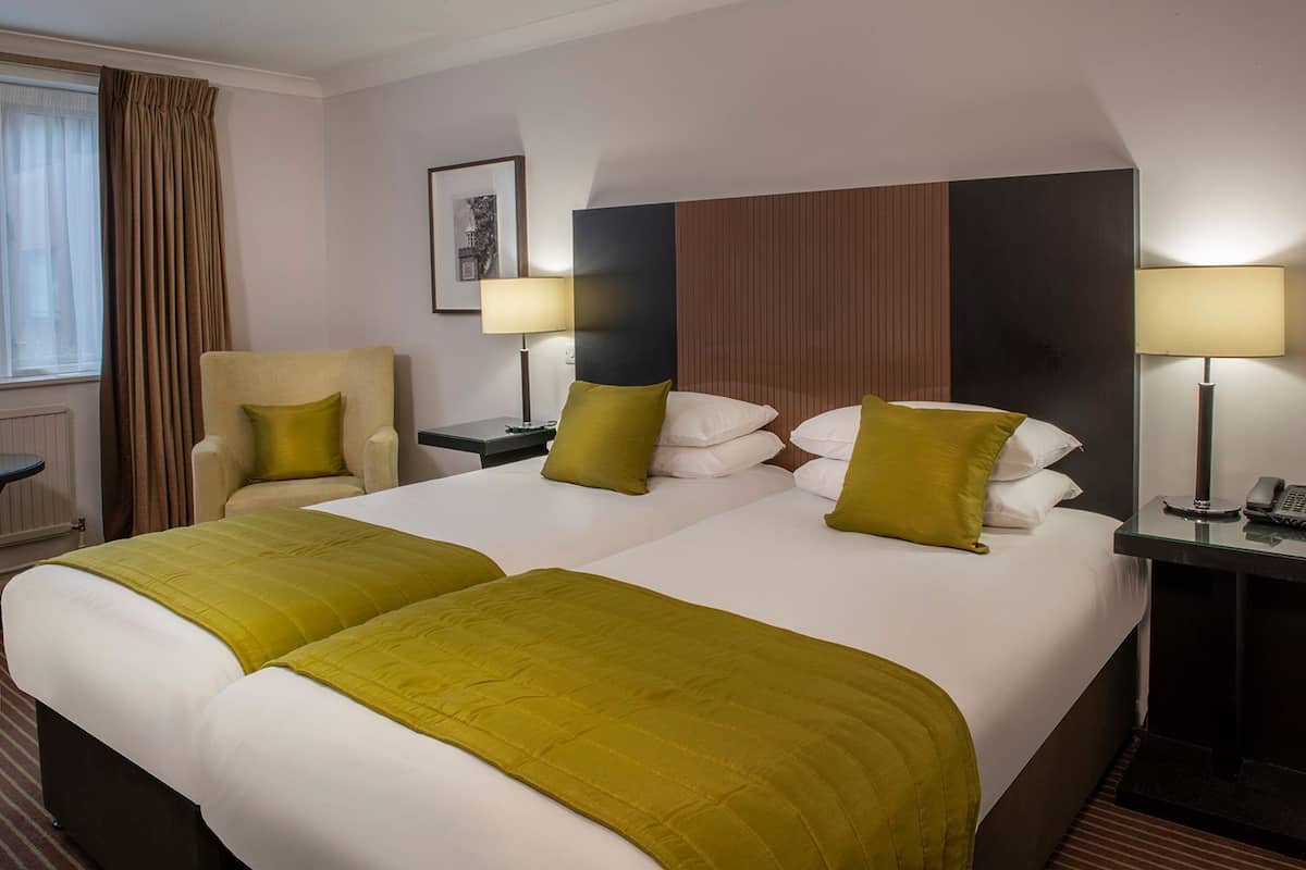 A hotel room with two beds, a chair, a desk, and a lamp.