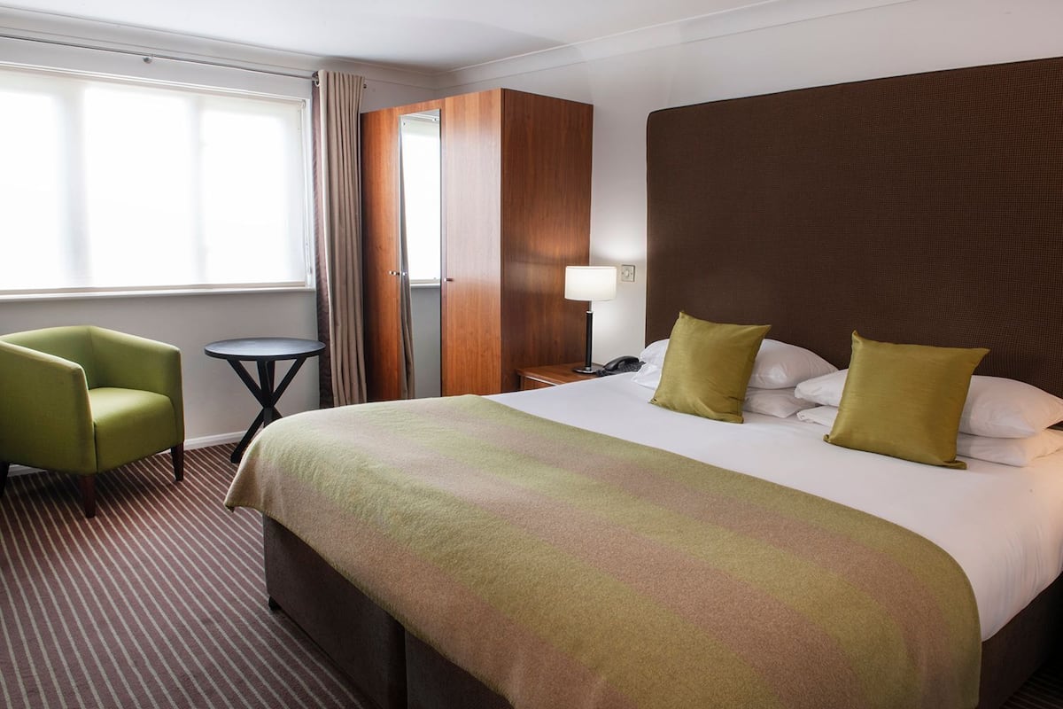 A hotel room with a large bed, a green armchair, a small round table, and a window with curtains.