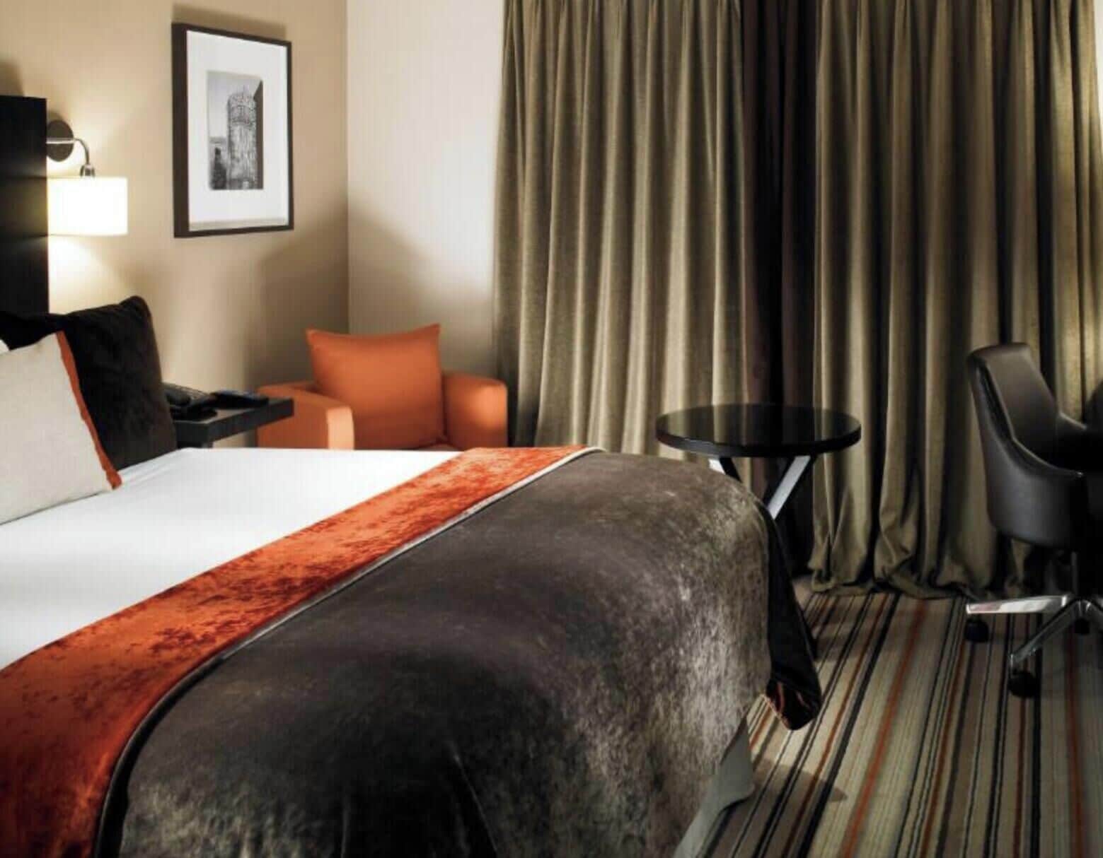 Quarto de hotel com cama, escrivaninha, cadeira, mesa pequena e um quadro na parede.