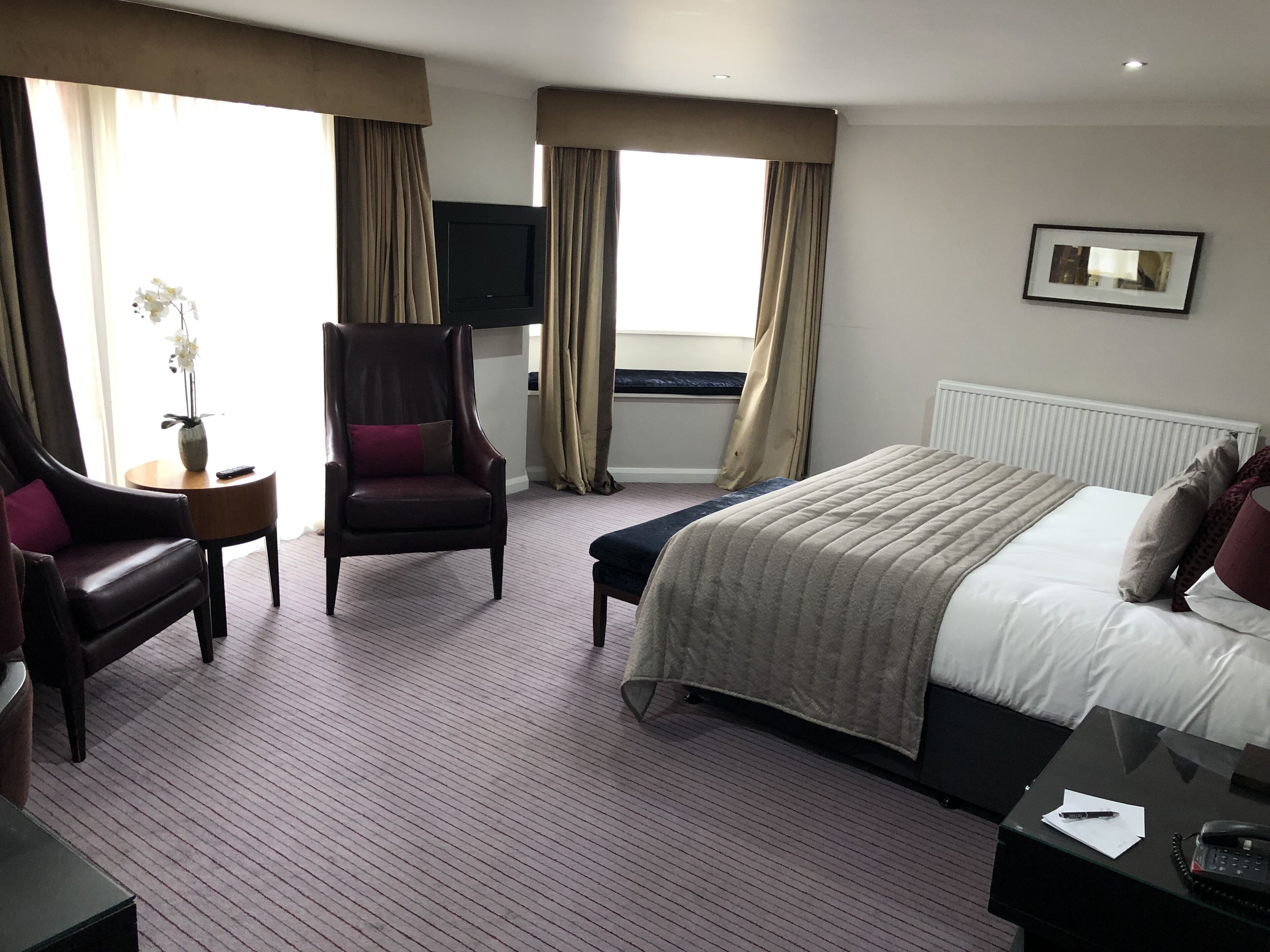 Una habitación de hotel con cama, una silla, una mesita con un jarrón y un televisor en la pared.