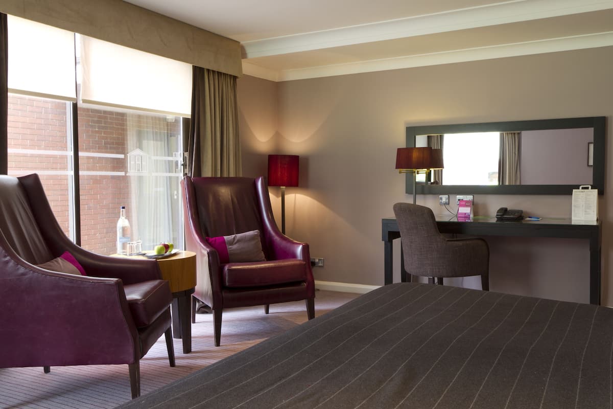 A hotel room with a large bed, two armchairs, a desk with a computer, a lamp, and a window with curtains.