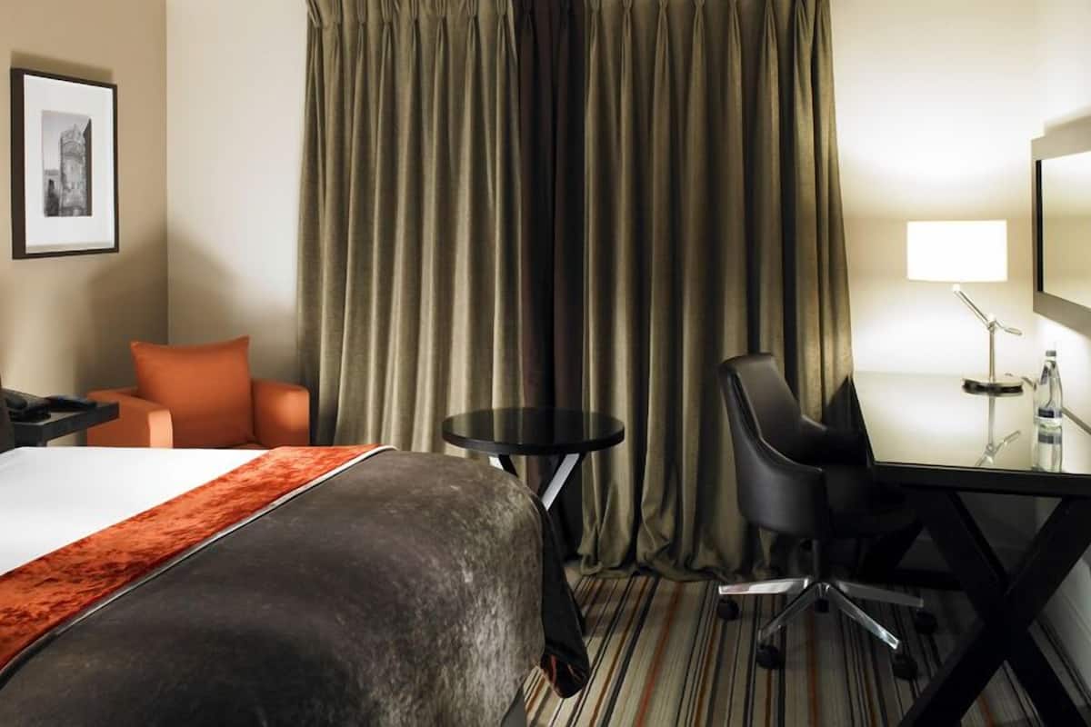 A hotel room with a bed, a desk, a chair, a small table, and a large window with curtains.