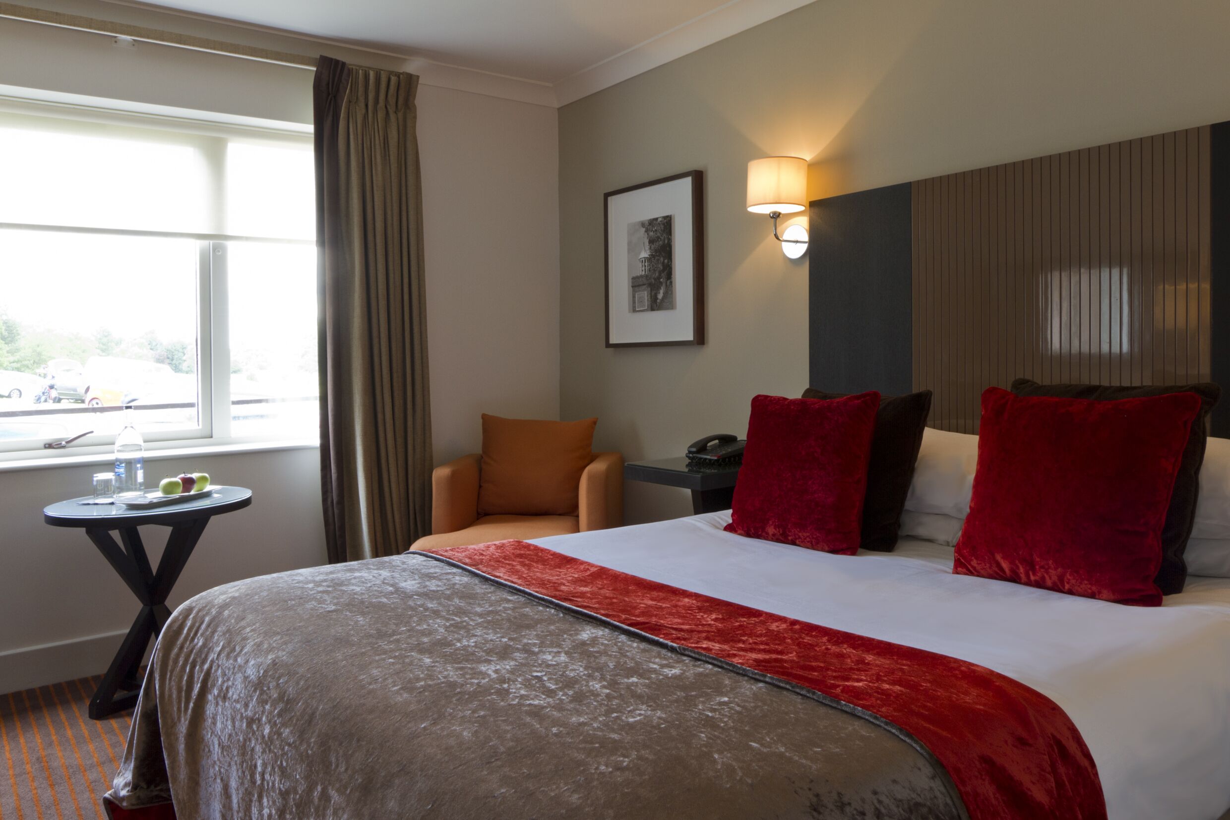 Ein Hotelzimmer mit einem großen Bett, einem kleinen Tisch mit Wasserflasche und Obst, einem Sessel und einem Fenster mit Vorhängen.