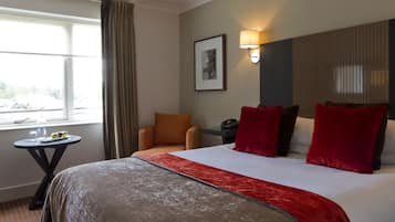 Ein Hotelzimmer mit einem großen Bett, einem kleinen Tisch mit Wasserflasche und Obst, einem Sessel und einem Fenster mit Vorhängen.